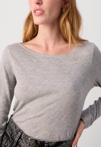 Sivi dugih rukava majica izrađena od mekog materijala s širokim dekolteom. Model nosi traperice, prikazujući ležeran stil.