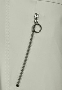 Diagonal silverdragkedja med en cirkulär dragflik på ljusbeige tyg med synlig sömnad.