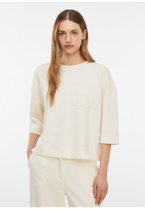 Junge Frau mit langen Haaren, die ein lockeres, cremefarbenes Oberteil mit Brusttasche und passende Hose trägt, steht mit den Händen in den Taschen.