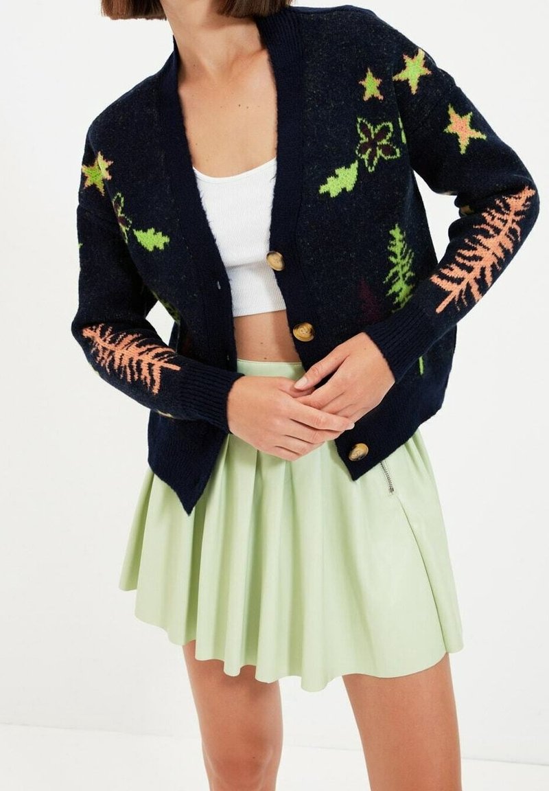 Femme portant un cardigan bleu marine boutonné avec des motifs de feuilles vertes et pêche, un haut court blanc et une jupe plissée vert pâle.