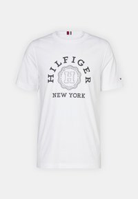 Tricou alb din bumbac, cu guler rotund, mâneci scurte și un grafic circular cu textul "HILFIGER" și "NEW YORK" în albastru închis.