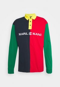 Pitkähihainen polo paita, jossa on väri-block -suunnittelu tummansinisessä, punaisessa, vihreässä ja keltaisessa, nappikauluksella ja valkoisella logo-yksityiskohdalla.