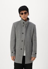 Cappotto grigio in misto lana tessuto con colletto alto, bottoni frontali e tasche laterali. Modello con occhiali da sole e una camicia bianca sotto.