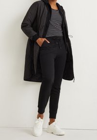 Cappotto lungo imbottito nero con cappuccio, indossato sopra una maglia a righe bianche e nere. Abbinato a pantaloni jogger neri e sneakers bianche.