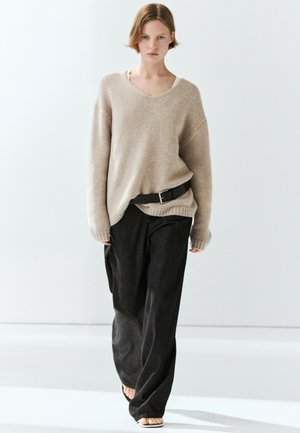 Kvinde går iført en løs beige strikket sweater, sorte bukser med vide ben og sort bælte samt åbne sandaler mod en ensartet lys baggrund.