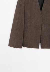 Bruine wollen blazer met zichtbare stiksels, licht uitlopende zoom en open voorkant tegen een effen witte achtergrond.