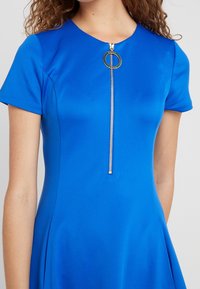 Blått figursytt klänning med korta ärmar, med en dragkedja framtill och en cirkulär ring att dra i. Slät tyg med en lätt lyster.