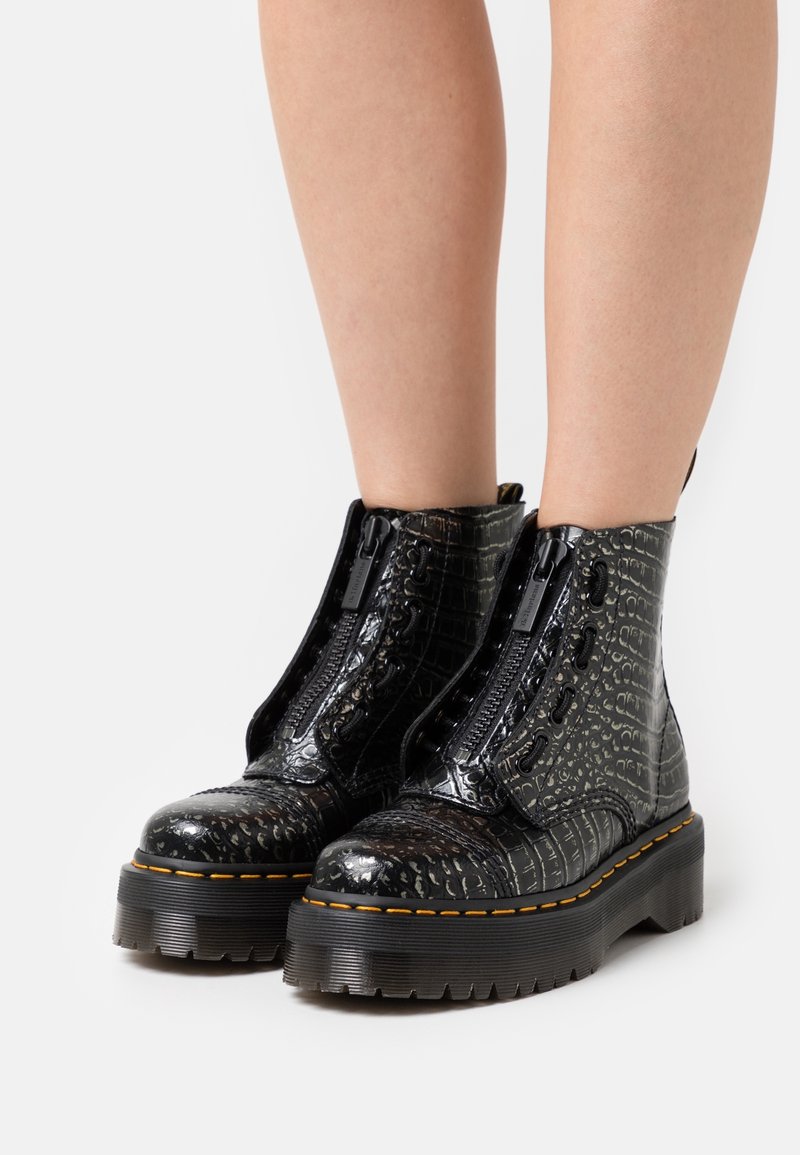 Bottes plateformes noires montant à la cheville avec texture crocodile, fermetures éclair avant et coutures jaunes, portées sur jambes nues sur fond blanc.