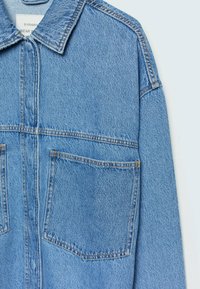 Světle modrá džínová bunda s límečkem, předním zipem a dvěma velkými náprsními kapsami s prošitým detailem.