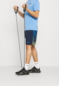 Camisa azul de manga corta, pantalones cortos azul marino con logo de Nike en degradado verde, zapatillas de entrenamiento negras, sosteniendo bandas de ejercicio con mangos negros.