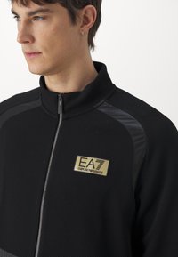 Felpa nera con zip, caratterizzata da un tessuto testurizzato, dettagli lucidi e una patch con il logo dorato "EA7 Emporio Armani."