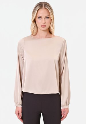 Giovane donna con capelli biondi che indossa una camicia beige a maniche lunghe e pantaloni neri, in piedi davanti a uno sfondo bianco neutro.