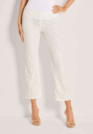 Vrouw draagt witte kanten wijde broek en beige open teen hakken, staand met één hand op de heup en de andere ontspannen.