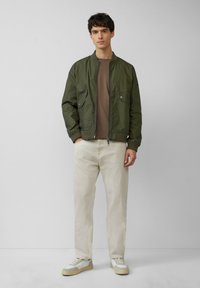 Olijfgroene bomberjack met rits aan de voorkant, twee voorzakken, gecombineerd met een bruine ronde hals T-shirt, beige broek en lichte sneakers.