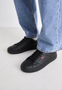 Fekete cipőfűzős sneaker, sima szintetikus anyagból, perforált részletekkel és piros logóval, világoskék farmernadrággal párosítva.