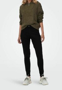 Zielona oliwkowa sweter z dzianiny o wysokim dekolcie i srebrnym zamkiem, połączony z czarnymi spodniami skinny i białymi sneakersami.