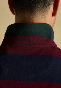 Marokānu un tumši zilu svītrotu polo krekls ar zaļu ribotu apkakli. Zīmols "JOULES" ir izšūts apkakles aizmugurē.