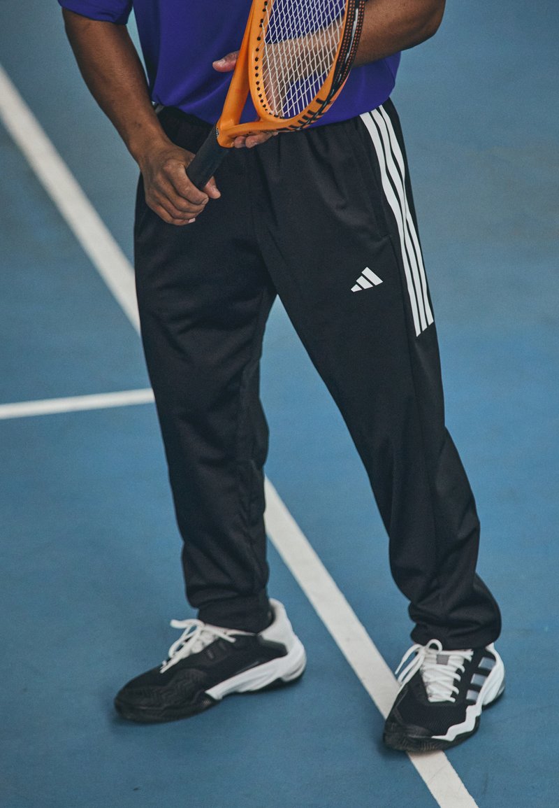 Svart sportbyxor med vita Adidas-stripes, kombinerat med svart och vita sneakers. Hållaren har ett tennisracket, bakgrunden är en blå tennisbana.