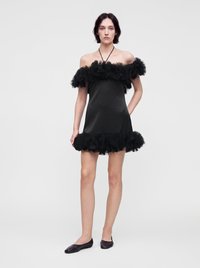 Γυναίκα με κοντό μαύρο φόρεμα χωρίς ώμους με βολάν στο πάνω μέρος και στο τελείωμα, συνδυασμένο με μαύρες μπαλαρίνες, στέκεται μπροστά σε ένα απλό φόντο.