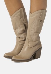 Stivali da cowboy in pelle beige con cuciture decorative, punta appuntita e tacco a blocco. Presentano una cerniera laterale per una facile indossabilità.