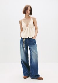 Kvinna med kort brunt hår klädd i en beige stickad ärmlös topp, vida blå jeans, bruna skor, ståendes mot vit bakgrund.
