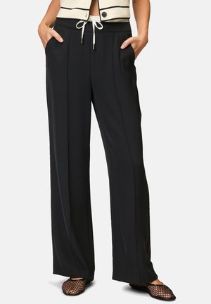 Persoon draagt zwarte wijde broek met plooien aan de voorkant, elastische tailleband met witte koord, handen in de zakken en zwarte visnet ballerina's.