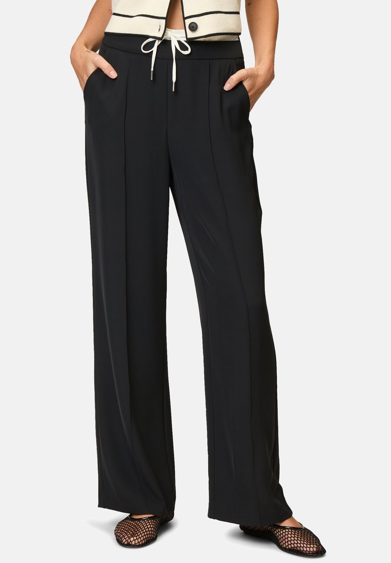 Personne portant un pantalon noir à jambes larges avec des plis devant, taille élastique avec cordon blanc, mains dans les poches et ballerines résille noires.