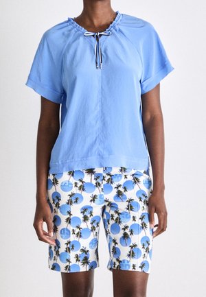 Jasnoniebieska bluzka z krótkim rękawem z wiązaniem z przodu, zestawiona z białymi szortami z nadrukiem niebieskich kół i palm.
