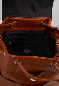 Borsa a secchiello in pelle marrone con finitura texturizzata, dotata di un interno nero, chiusura con coulisse e una tasca con zip. Etichetta del brand visibile all'interno.