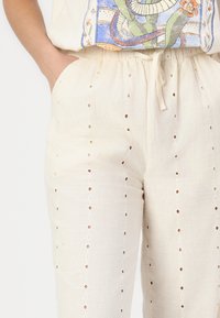 Pantalons en lin couleur beige clair avec des découpes en œillets verticales, une taille à cordon à nouer et une coupe décontractée.