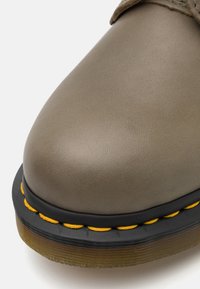 Zapato de cuero marrón con punta redonda, suela de goma y borde negro. Presenta un pespunte amarillo a lo largo de la suela y una superficie texturizada.