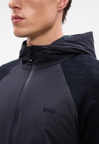 Casaco preto com capuz e fecho de zip, tecido texturizado nos ombros e logotipo em relevo no peito. Acabamento suave com detalhes escuros contrastantes.