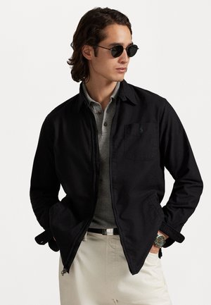 Zwarte zip-up jas met een puntige kraag, enkele borstzak, gedragen over een grijze polo. Lichtgekleurde broek en ronde zonnebril maken de look compleet.