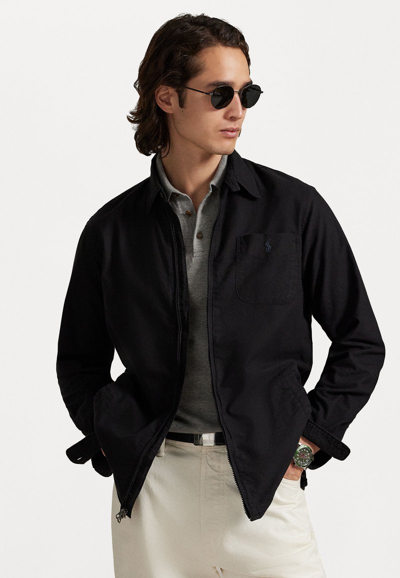 Black zip-up jacket with a pointed collar, single chest pocket, worn over a gray polo. Light-colored pants and round sunglasses complete the look.