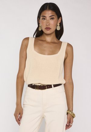 Donna che indossa un top beige senza maniche lavorato a maglia, pantaloni crema a vita alta con cintura scura, grandi orecchini e un bracciale spesso su sfondo semplice.