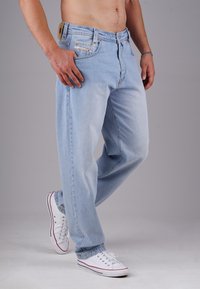 Mann in hellblauen Jeans und weißen Sneakers, der mit einer Hand in der Tasche vor einem grauen Hintergrund steht.