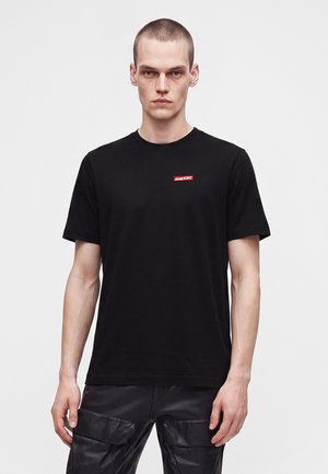 Mand iført en simpel sort t-shirt med korte ærmer med et lille rødt logo på brystet og sorte cargobukser mod en ensartet baggrund.