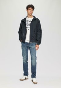 Marineblauwe bomberjack met een rits aan de voorkant, gecombineerd met een witte graphic t-shirt, blauwe jeans en witte sneakers met bruine accenten.