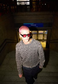 Siva pletjena pulover s frayed roba, v kombinaciji s črnimi hlačami. Okrogla, prevelika rdeča sončna očala. Model hodi po kamnitih stopnicah.