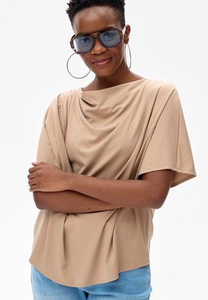 Kobieta w dużych niebieskich okularach przeciwsłonecznych, dużych kolczykach kołach, beżowej zwiewnej bluzce i jasnoniebieskich dżinsach, z założonymi ramionami, lekko się uśmiecha.