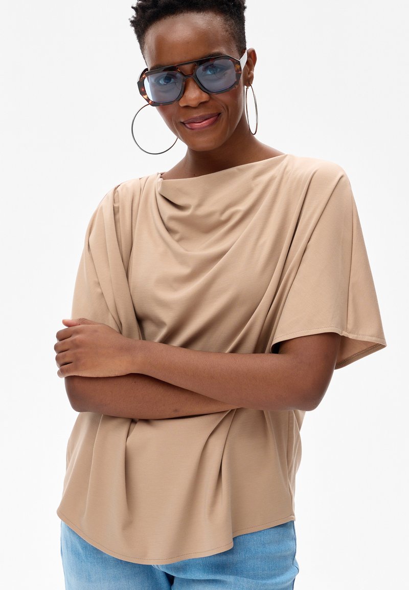 Kobieta w dużych niebieskich okularach przeciwsłonecznych, dużych kolczykach kołach, beżowej zwiewnej bluzce i jasnoniebieskich dżinsach, z założonymi ramionami, lekko się uśmiecha.