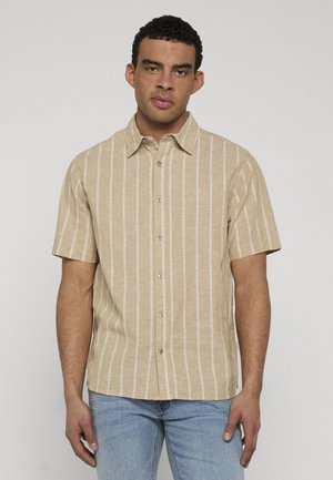 Mann iført beige kortermet skjorte med hvite vertikale striper og lyseblå jeans, stående mot en ensfarget bakgrunn.