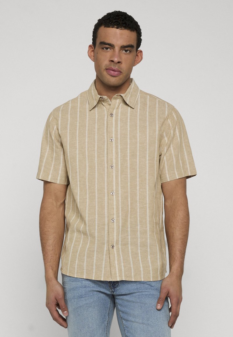 Homme portant une chemise beige à manches courtes avec des rayures blanches verticales et un jean bleu clair, debout devant un fond uni.