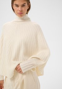 Kobieta w kremowym swetrze z prążkowanym golfem i luźnymi rękawami, włożona w wysokie, kremowe spodnie na jednolitym jasnym tle.