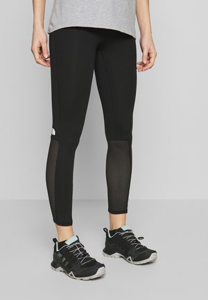 Person iført sorte leggings med meshpaneler på læggene og sorte trail løbesko med lyseblå accenter.