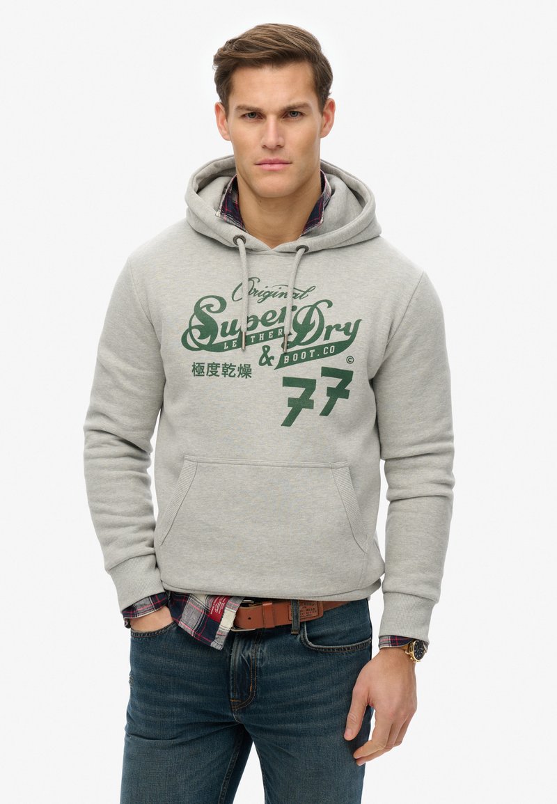 Grå hættebluse med kengurulomme, med grønnt trykdesign og nummer "77." Båret over en ternet skjorte, med jeans og et bælte.