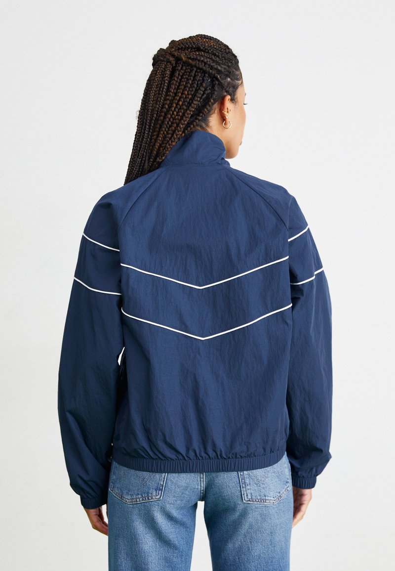 Personne avec des cheveux tressés portant une veste coupe-vent bleu marine avec des rayures blanches en chevron, associée à un jean bleu clair, de dos.