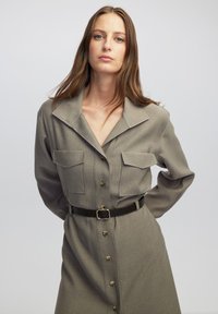 Khaki-kleurige shirtjurk met een kraag, dubbele borstzakken, knoopsluiting aan de voorkant en een ceintuur in de taille. De stof heeft een subtiele textuur.