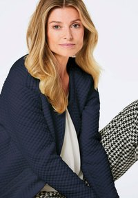 Marineblauer gesteppter Mantel mit Reißverschluss, kombiniert mit schwarz-weißen Hahnentrittmuster-Hosen. Das Model hat lange, wellige Haare und ein neutrales Oberteil.