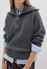 Suéter gris de punto con cuello con cremallera, que presenta un dobladillo y puños acanalados. Debajo, se vislumbran los puños de una camisa de rayas azul claro.